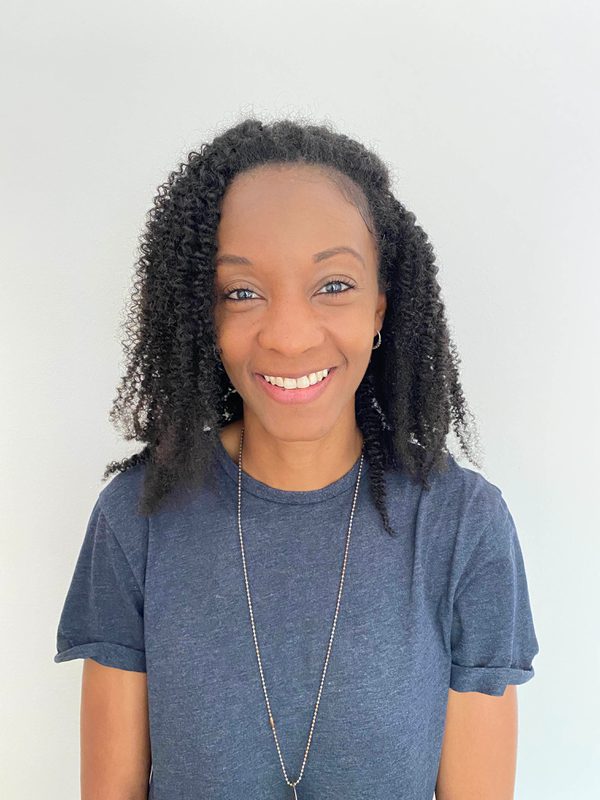 A woman wearing a blue short sleeved t-shirt and a long necklace smiles at the camera, there is a cream background behind her.