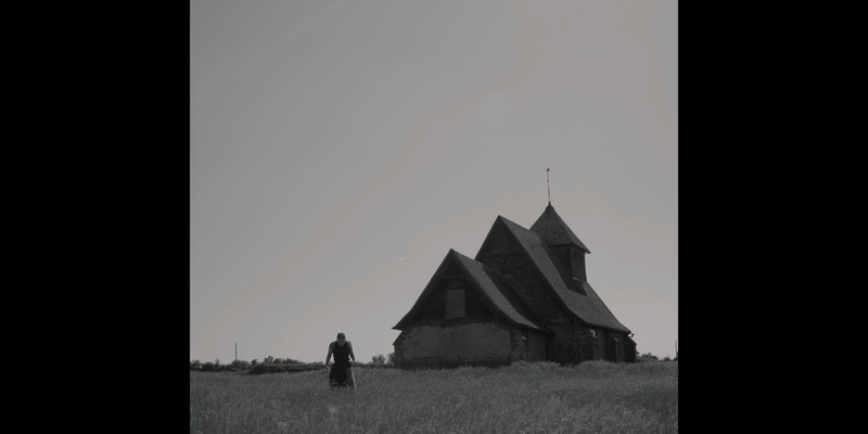 A black and white photo with a large house on the right hand side and a woman can be seen on the left in the long grass, she is looking down