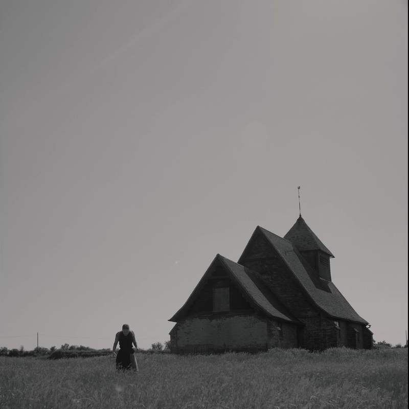 A black and white photo with a large house on the right hand side and a woman can be seen on the left in the long grass, she is looking down