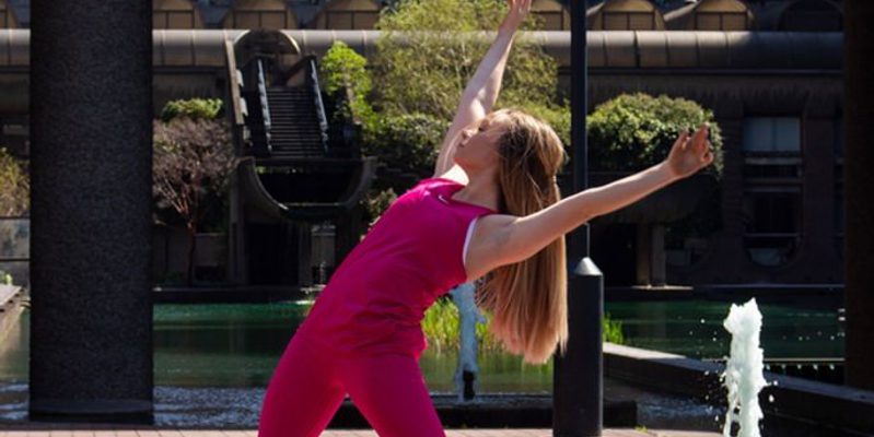 Image of Ellie dancing centrally in the camera. Behind Ellie is a water feature and a small tree. Ellie is wearing all pink clothes and has long blonde hair.
