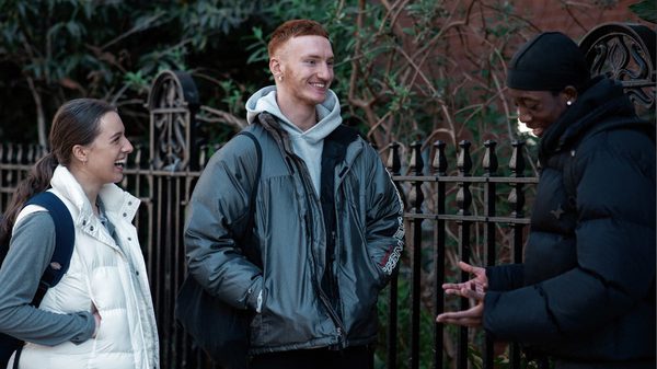 Three LCDS students stand outside The Place, there is a metal fence and trees behind them, one student is making the other two laugh