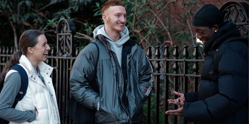 Three LCDS students stand outside The Place, there is a metal fence and trees behind them, one student is making the other two laugh