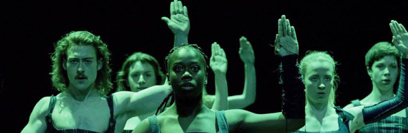 Five dancers wearing corsets and in deep green lighting. Their left arm raised in the air, palm facing forwards, right arm over their groin.