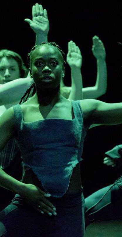Five dancers wearing corsets and in deep green lighting. Their left arm raised in the air, palm facing forwards, right arm over their groin.