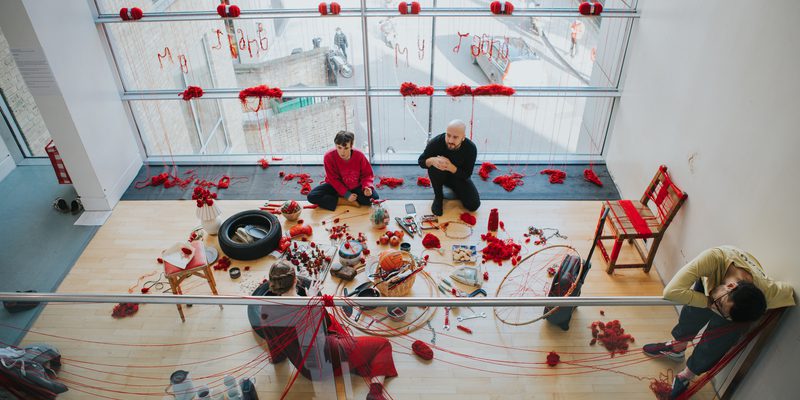 A mezzanine level floor with a wall of windows captured by an image taken slightly from above. Red yarn has been spread around the entire space, hanging over the balcony, across the widows and forming small piles on the floor. There is a large assortment of household items and trinkets that have been wrapped in red yarn. Two people sit around these items, face to face with another person, whilst a final person stands up leaning their body over the balcony.