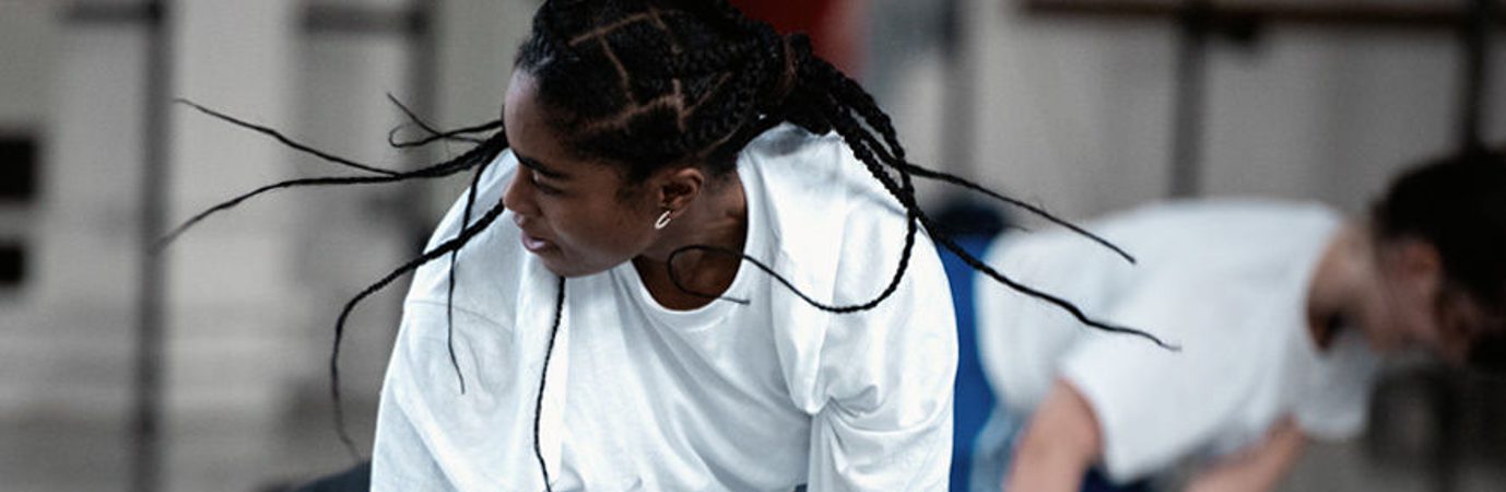 A person wearing a white long sleeved top in our studio. They are on the floor with their arms are stretched to the floor and their head is looking to the right, which makes their hair braids fly out