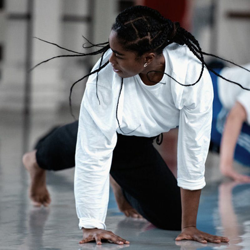 A person wearing a white long sleeved top in our studio. They are on the floor with their arms are stretched to the floor and their head is looking to the right, which makes their hair braids fly out