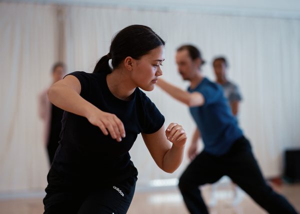 A person in the forefront of the image is wearing a black t-shirt and trousers. They are standing, knees bent, and their body is twisted as they look to the left towards the floor. Their right arm reaches forward with a bent elbow, palm facing the floor. The left arm is bent by the side of their body. In the background here are three other dancers in the studio