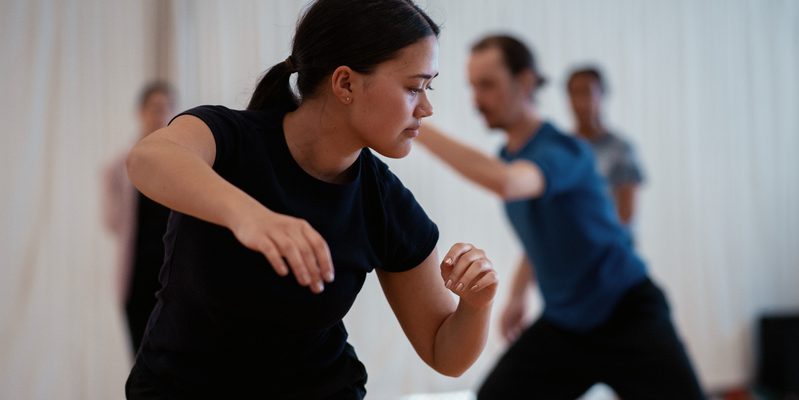 A person in the forefront of the image is wearing a black t-shirt and trousers. They are standing, knees bent, and their body is twisted as they look to the left towards the floor. Their right arm reaches forward with a bent elbow, palm facing the floor. The left arm is bent by the side of their body. In the background here are three other dancers in the studio