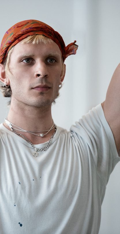 In a dance studio, a dancer in a white t-shirt with an orange scarf around their head stands with both arms reaching upwards, elbows slightly bent. A dancer in the background stands in the same position, arms up and elbows bent with palms facing towards each other. They wear a black t-shirt.