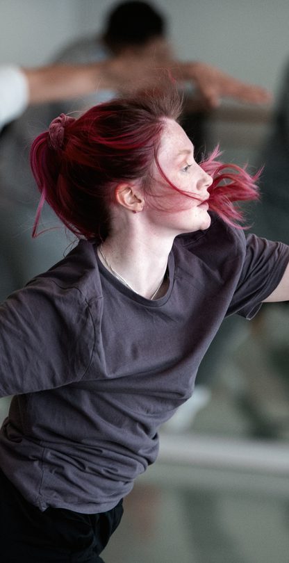 Several dancers are in the same position in one of our studios. The dancer in the foreground has pink hair and wears a dark grey t-shirt and black bottoms. They a lunging forward in a slight twist, looking towards their left arm which is fully stretched out in front of them, parallel to the floor. The other arm is lifted away from the body and is directed towards the floor.