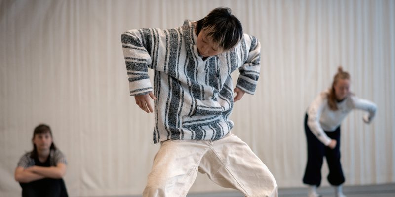In the studio, a dancer in black bottoms and a light grey t-shirt sits upright with both knees bent and their forearms wrapped around their knees. They are looking at a dancer in the foreground who is wearing white trousers and a light striped hooded top. Their knees are bent and their arms are bent at a right angle towards the floor. They are looking down. Behind and to the right of them, a final dancer in a white top and black bottoms leans forward and holds their arms away from their body with elbows bent.