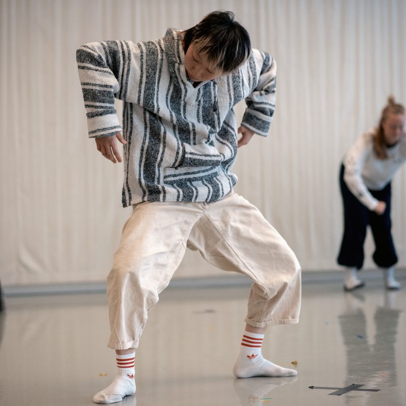 In the studio, a dancer in black bottoms and a light grey t-shirt sits upright with both knees bent and their forearms wrapped around their knees. They are looking at a dancer in the foreground who is wearing white trousers and a light striped hooded top. Their knees are bent and their arms are bent at a right angle towards the floor. They are looking down. Behind and to the right of them, a final dancer in a white top and black bottoms leans forward and holds their arms away from their body with elbows bent.