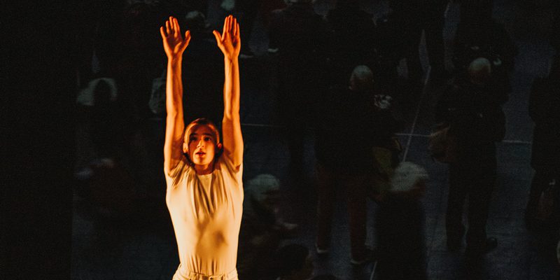 Two dancers, one rolling across the floor, the other jumping over them with their arm stretched to the ceiling and knees bent. They are both wearing white.