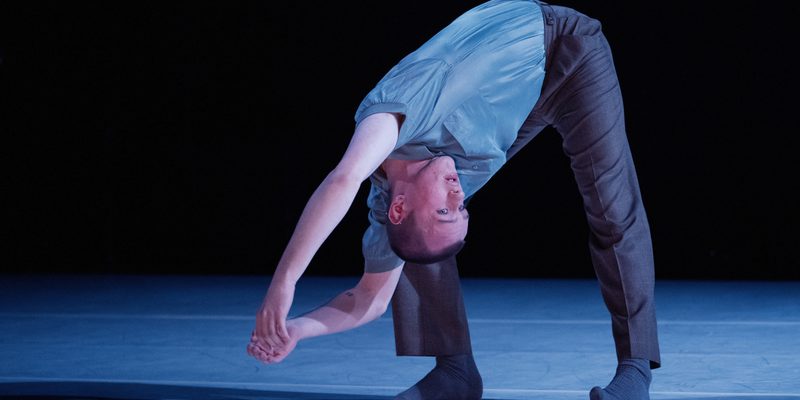 A dancer wearing a blue t-shirt, grey trousers and dark socks is lit by a blue wash of lighting on stage. Their left leg straight and right leg slightly bent, they hang lean their torso over so it is directed diagonally towards the floor. Their body is twisted to face stage left, and their arms are outstretched above and behind their head. The top of their head is facing the floor, and they crane their neck forward staring into the distance.