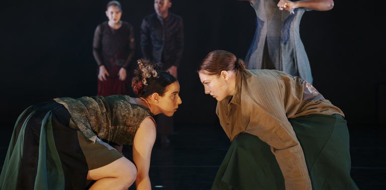 Two dancers crouch face-to-face in the foreground of a dark stage, while other performers pose behind them in layered earth-toned costumes under warm theatrical lighting.