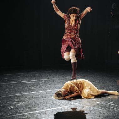 A dancer leaps high above another performer curled on the floor, both lit by a warm spotlight on a dark stage.