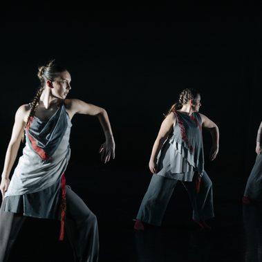 Three dancers in gray layered costumes pose on a dark stage, each turning to the side with one arm bent under focused theatrical lighting.