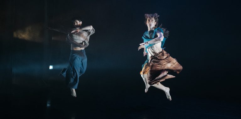 Two dancers leap across a dark stage, arms bent in motion, wearing loose layered costumes under cool blue theatrical lighting.