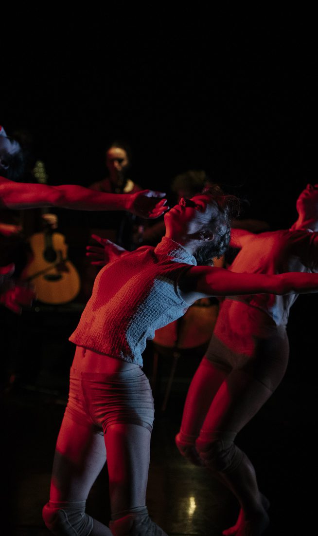 Four dancers in light costumes move with outstretched arms under red stage lighting. Behind them, an ensamble of musicians is dimly visible in the dark background.