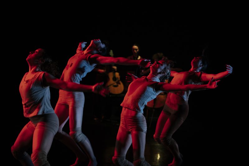 Four dancers in light costumes move with outstretched arms under red stage lighting. Behind them, an ensamble of musicians is dimly visible in the dark background.