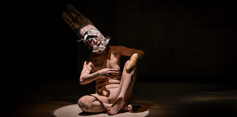 A performer sits in a spotlight on a dark stage wearing a tall paper headdress and white face makeup. Their body is folded into a contorted pose, with one leg raised and an arm crossing their torso.