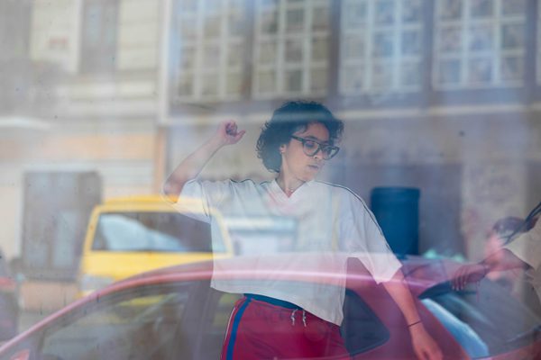 A person wearing glasses, a white T-shirt, and red pants is captured mid-movement behind a window, with one arm raised as if dancing. Reflections of cars and buildings on the glass partially overlap their figure, creating layered views of the street and the indoor space.