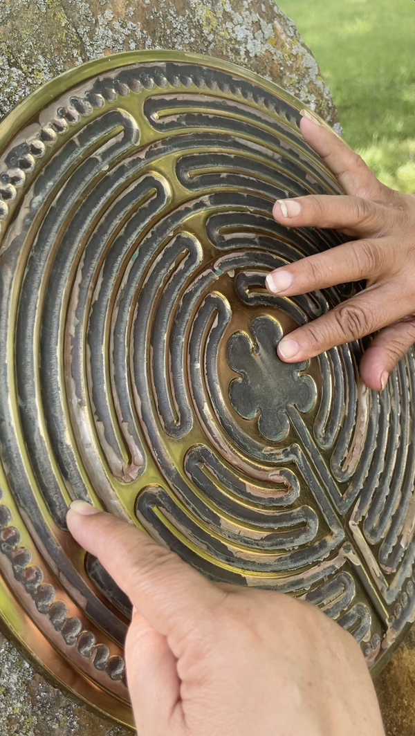 Two hands trace the raised pathways of a circular bronze labyrinth mounted against a tree trunk outdoors. The close-up view highlights the textured metal surface and the fingers following the winding pattern toward the centre.