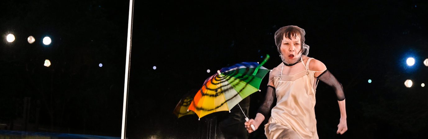 A performer in a cream dress and mesh head covering runs across a stage holding a brightly coloured umbrella. Reflections in a mirrored wall behind them echo the movement, while stage lights illuminate the dark performance space.