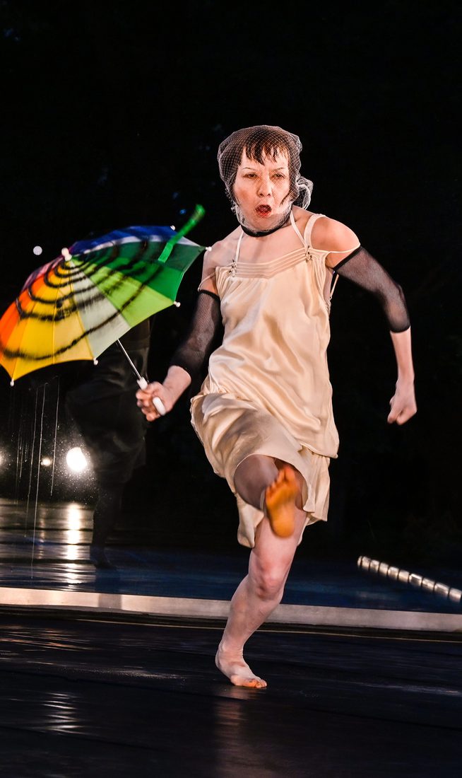 A performer in a cream dress and mesh head covering runs across a stage holding a brightly coloured umbrella. Reflections in a mirrored wall behind them echo the movement, while stage lights illuminate the dark performance space.
