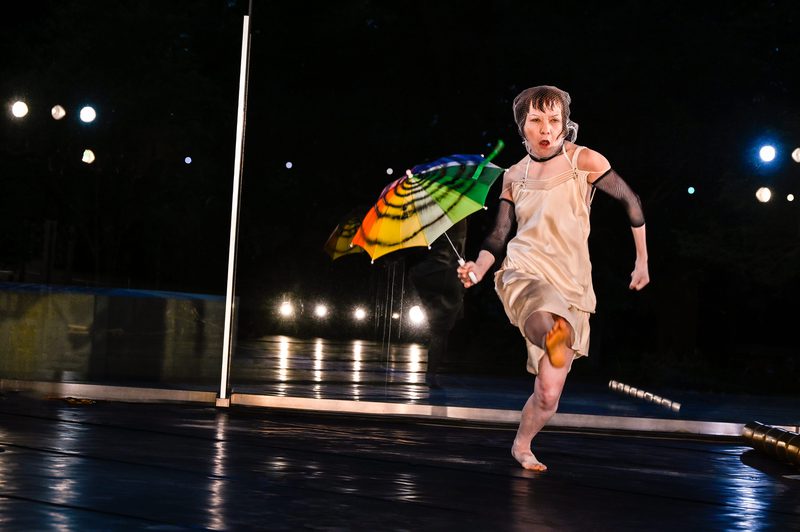 A performer in a cream dress and mesh head covering runs across a stage holding a brightly coloured umbrella. Reflections in a mirrored wall behind them echo the movement, while stage lights illuminate the dark performance space.
