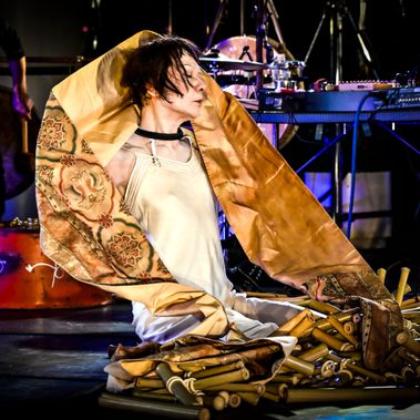 A performer sits on a stage draped in patterned fabric, surrounded by bamboo tubes and musical equipment. The fabric frames their head and shoulders, creating a sculptural silhouette.