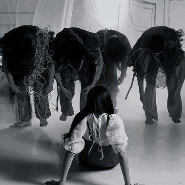 A person with long dark hair sits on the floor facing four figures dressed in dark, tattered clothing. The four figures stand bent forward with their heads lowered, forming a line in front of sheer draped fabric. One figure has a bundle of rope or cords hanging over their shoulder. The image is monochrome, with a sparse stage-like setting and a strong contrast between the seated figure and the group.
