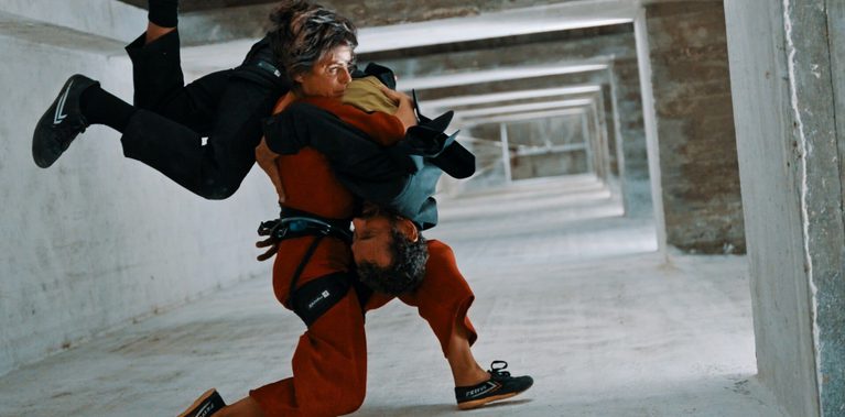 Two performers are captured in a dynamic acrobatic movement inside a stark concrete structure. One performer, wearing a rust-coloured overall, crouches low while supporting a second performer dressed in black, whose body is inverted and wrapped around them. The pair hold each other tightly as they balance, creating a sculptural, intertwined shape. The setting is an empty, unfinished corridor with repeating concrete frames receding into the distance.
