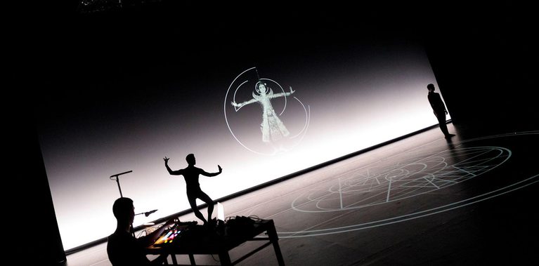 A wide-angle, high-contrast view of a stage featuring layered projections and silhouettes. Centre stage, a dancer appears as a sharp silhouette against a bright horizontal light band. The floor is covered in a projected geometric grid of circles, while the back wall shows a circular projection of a figure in ornate dress. Above, a large, curved sheet of shimmering silver material hangs from the ceiling.