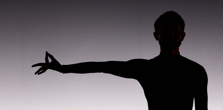 A close-up, high-contrast silhouette of a performer's upper body against a bright, gradient white background. The dancer is facing forward, with one arm extended straight out to the side. Their hand is held in a precise mudra-like gesture, with the thumb and forefinger touching to form a small circle while the other fingers are fanned out.