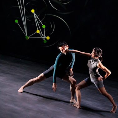 Two dancers perform on a dark stage in front of a large digital projection. The projection consists of yellow and green glowing dots connected by thin white lines, resembling a constellation map against a backdrop of faint, rippling circles. In the foreground, a dancer in a teal long-sleeved top and black shorts lunges deeply to the left. Beside them, a dancer in a grey sleeveless top and shorts stands in a wide stance, extending one arm across the other's face in a sharp, linear gesture.
