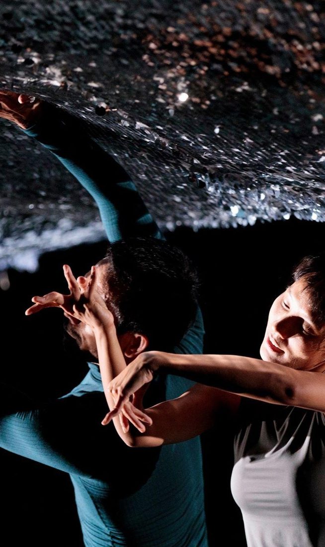 Two performers are captured in a tight, layered composition beneath a low, textured ceiling covered in shimmering silver sequins. In the foreground, a dancer in a grey sleeveless top leans forward with their arms held in a fluid, bent pose. Behind them, a second performer in a teal long-sleeved top reaches one hand up to touch the sparkling surface while their other hand is held near their face in an angular gesture.