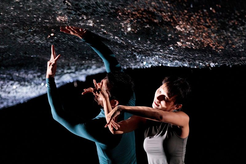 Two performers are captured in a tight, layered composition beneath a low, textured ceiling covered in shimmering silver sequins. In the foreground, a dancer in a grey sleeveless top leans forward with their arms held in a fluid, bent pose. Behind them, a second performer in a teal long-sleeved top reaches one hand up to touch the sparkling surface while their other hand is held near their face in an angular gesture.