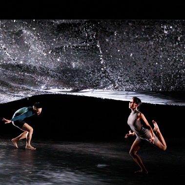 A wide shot of two performers on a dark stage beneath a vast, undulating canopy of shimmering silver sequins. One dancer in a teal top is crouched low to the ground in a deep lunge, while the other in a grey top moves with a leg raised behind them. The floor is dappled with reflected light from the ceiling.