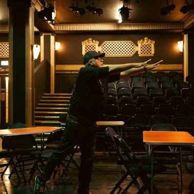 A man dressed in black with back cap reaches both arms to one side. His is in a dark theatrical space with dim lighting, tables spread around the room and ranked empty seating in the background.