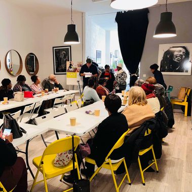 A room with tables and chairs in a U shape. Many adults sit with drinks and snacks before them, listening attentively to a man who is reading from papers at the front of the room.