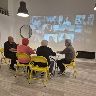 Four adults sit on yellow chairs at a table before an image of many students beamed onto the wall. The live and video participants are looking at each other.