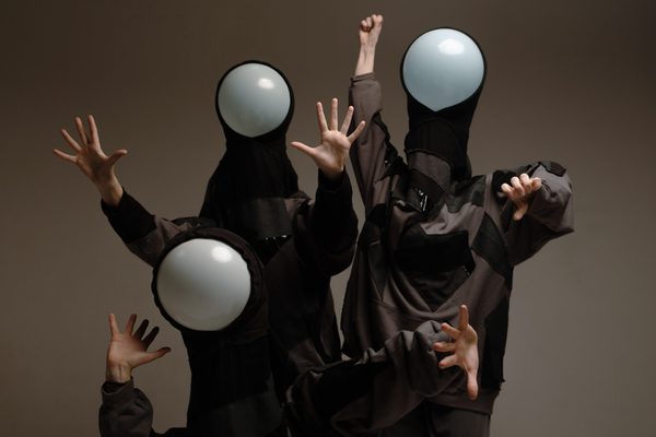 Three performers pose dynamically against a neutral gray background. They wear dark, oversized streetwear with patchwork textures and black hoods that feature large, opaque white orbs in place of faces. Their hands are raised in various expressive gestures, including open palms and a raised fist.