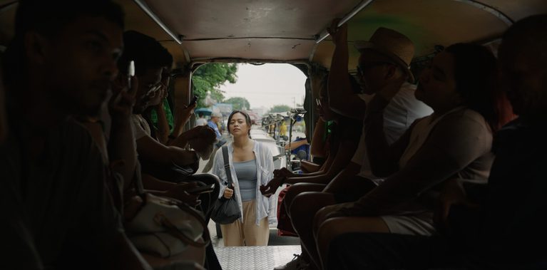 View from inside a crowded vehicle looking toward the open rear entrance. Passengers sit along both sides, many looking at their phones. A woman stands outside at the doorway, facing in, with a bag over one shoulder. A busy street is visible behind her.