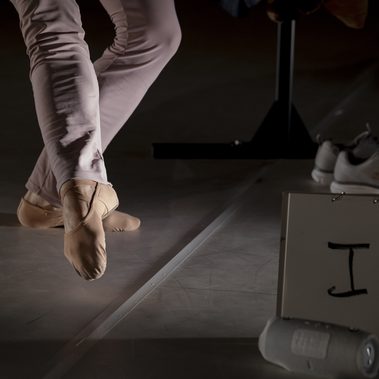 Close-up of a performer’s feet in soft dance shoes crossing on tiptoe under stage lighting. In the foreground, a white sign with the handwritten words ‘I was’ is partially visible beside a plastic bottle, while a pair of trainers can be seen in the background.