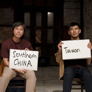 Two performers sit on chairs in a theatre space, each holding a white sign with handwritten text. The sign on the left reads ‘Southern China’ and the sign on the right reads ‘Taiwan’. Both look off to one side while seated against a backdrop of dark wooden doors and weathered walls.
