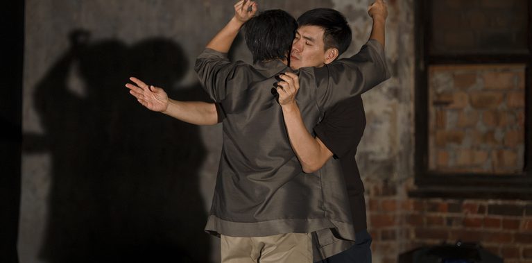 Two performers stand close together, holding each other while extending their arms in different directions. One performer faces the camera and the other faces away. Their shadows are projected onto the wall behind them. The image is set in a theatre space with worn plaster walls and exposed brick.