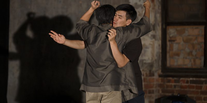 Two performers stand close together, holding each other while extending their arms in different directions. One performer faces the camera and the other faces away. Their shadows are projected onto the wall behind them. The image is set in a theatre space with worn plaster walls and exposed brick.