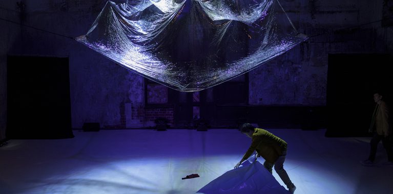 A performer unfolds a large sheet of translucent plastic on the floor of a dimly lit performance space. Above them, a suspended canopy of reflective plastic hangs from the ceiling, illuminated by blue and purple stage lighting. Another figure stands at the edge of the space, while the backdrop features weathered walls and dark architectural details.